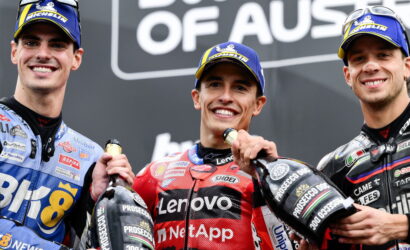 Three motorcycle racers on a winner's podium, smiling and holding bottles of champagne.