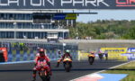 Motorcycle race on a track with riders in red and black racing suits, leading near the start/finish line under a large sponsor sign.
