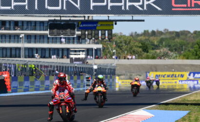 Motorcycle race on a track with riders in red and black racing suits, leading near the start/finish line under a large sponsor sign.