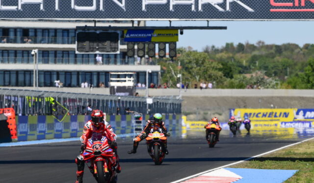 Motorcycle race on a track with riders in red and black racing suits, leading near the start/finish line under a large sponsor sign.