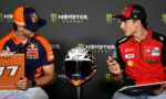 Two MotoGP riders in team gear sit and talk at a press event, with a racing helmet on a stand between them and clipboards in hand.