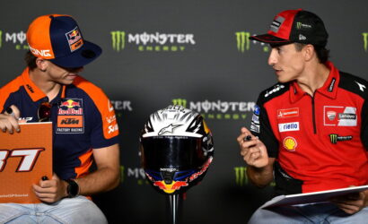 Two MotoGP riders in team gear sit and talk at a press event, with a racing helmet on a stand between them and clipboards in hand.