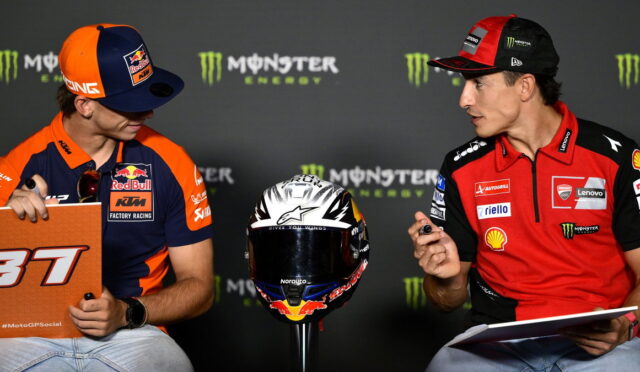 Two MotoGP riders in team gear sit and talk at a press event, with a racing helmet on a stand between them and clipboards in hand.