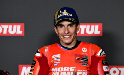 MotoGP rider in red race suit on podium, smiling at camera, wearing a Michelin cap with sponsor logos visible on suit and backdrop