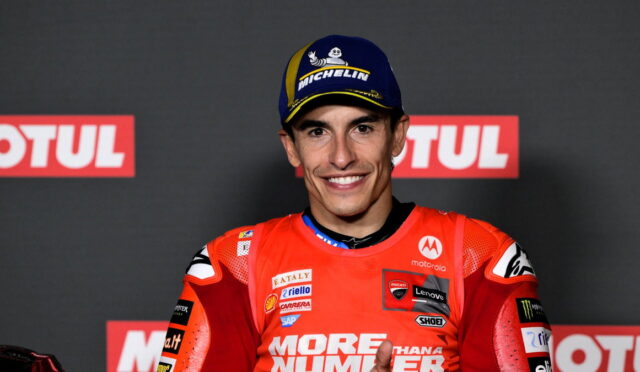 MotoGP rider in red race suit on podium, smiling at camera, wearing a Michelin cap with sponsor logos visible on suit and backdrop