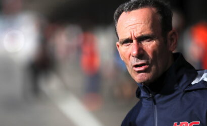 Close-up of a middle-aged man in a dark navy sports jacket outdoors, looking tense and focused, with a blurred background and a red HRC logo on his sleeve.