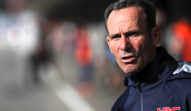 Close-up of a middle-aged man in a dark navy sports jacket outdoors, looking tense and focused, with a blurred background and a red HRC logo on his sleeve.