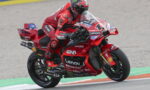 MotoGP rider in red Ducati livery leaning into a turn on a race track with sponsor decals visible in the background.