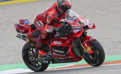 MotoGP rider in red Ducati livery leaning into a turn on a race track with sponsor decals visible in the background.