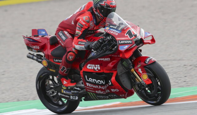 MotoGP rider in red Ducati livery leaning into a turn on a race track with sponsor decals visible in the background.