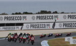 Motorcycle race on a track with riders leaning into a curve; large Prosecco DOC banners along the barrier in the background.