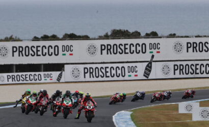 Motorcycle race on a track with riders leaning into a curve; large Prosecco DOC banners along the barrier in the background.