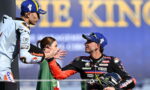 Two motorcycle racers shake hands on a podium after a race; one in a light-blue racing suit, the other in a black suit holding a helmet.
