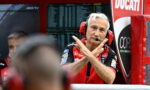 Ducati race team member in red shirt with headset and microphone, crossing his arms in an X gesture in the pit area.