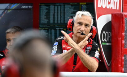 Ducati race team member in red shirt with headset and microphone, crossing his arms in an X gesture in the pit area.