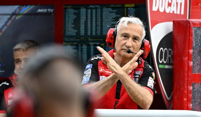 Ducati race team member in red shirt with headset and microphone, crossing his arms in an X gesture in the pit area.