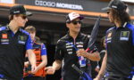 Three motorcycle racers in team uniforms chat in the pit area; the central rider holds a Red Bull umbrella and can.ensor