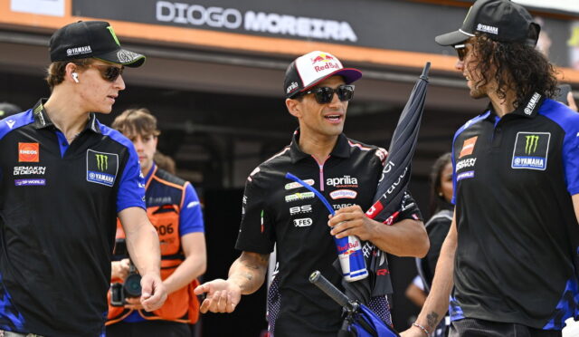 Three motorcycle racers in team uniforms chat in the pit area; the central rider holds a Red Bull umbrella and can.ensor
