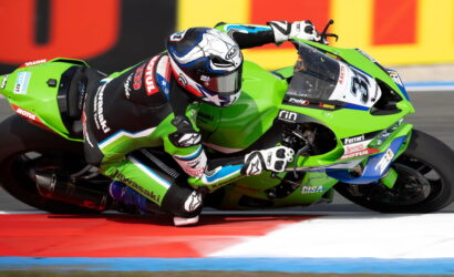 Rider on a green Kawasaki superbike leans into a turn on a race track, red-white-blue curbs visible in the foreground.