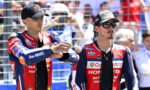 Two motorcycle racers in red‑blue Honda suits shake hands in front of a fenced crowd before a race, one wearing a black cap.
