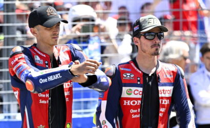 Two motorcycle racers in red‑blue Honda suits shake hands in front of a fenced crowd before a race, one wearing a black cap.