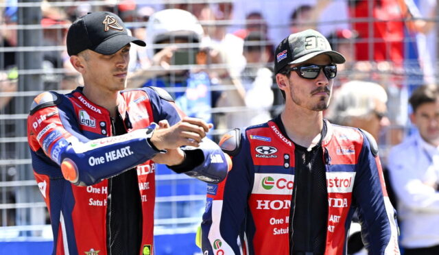 Two motorcycle racers in red‑blue Honda suits shake hands in front of a fenced crowd before a race, one wearing a black cap.