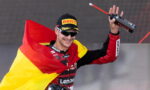 Male motorcycle racer on a podium, draped in the German flag, holding a trophy bottle and smiling in sunglasses and a Pirelli cap.