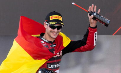 Male motorcycle racer on a podium, draped in the German flag, holding a trophy bottle and smiling in sunglasses and a Pirelli cap.