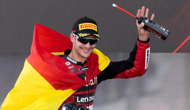 Male motorcycle racer on a podium, draped in the German flag, holding a trophy bottle and smiling in sunglasses and a Pirelli cap.