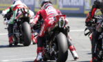 Motorcyclists racing on a track from behind, red-suited rider in the center leaning into a turn with a Monster energy exhaust visible on the bike's tail. The blur of rivals, tire marks, and asphalt emphasize speed and competition.