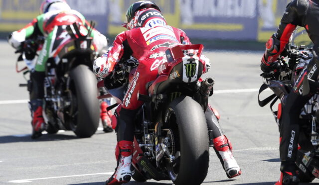 Motorcyclists racing on a track from behind, red-suited rider in the center leaning into a turn with a Monster energy exhaust visible on the bike's tail. The blur of rivals, tire marks, and asphalt emphasize speed and competition.