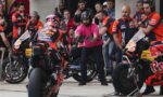 Pit lane scene with two Red Bull KTM motorcycles, crew in orange uniforms, and a man in pink signaling to riders.