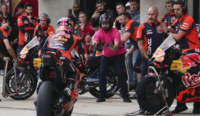 Pit lane scene with two Red Bull KTM motorcycles, crew in orange uniforms, and a man in pink signaling to riders.