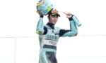 Racer in teal suit lifts helmet overhead in celebration on a podium, holding a water bottle in the other hand.