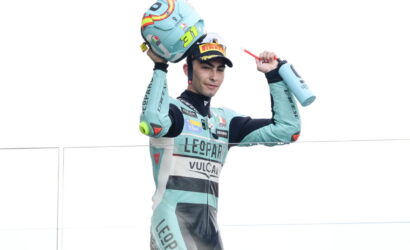 Racer in teal suit lifts helmet overhead in celebration on a podium, holding a water bottle in the other hand.