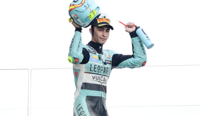 Racer in teal suit lifts helmet overhead in celebration on a podium, holding a water bottle in the other hand.