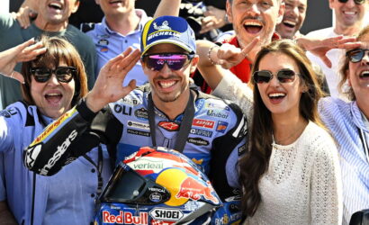 A crowd of fans cheer as a motorcycle racer in a blue racing suit poses with his Red Bull helmet.