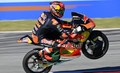 Rider on a Red Bull KTM motorcycle performs a wheelie on a racetrack, wearing a matching orange-black suit and helmet.