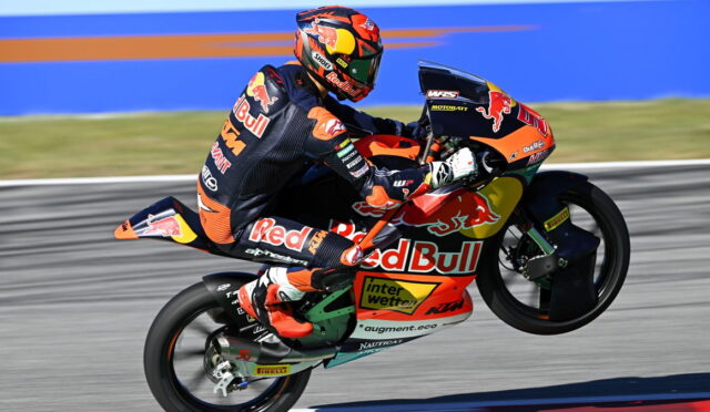 Rider on a Red Bull KTM motorcycle performs a wheelie on a racetrack, wearing a matching orange-black suit and helmet.