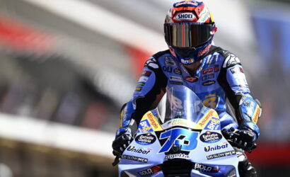 Motorcycle racer in a blue suit and red helmet speeds on a blue race bike at a track.