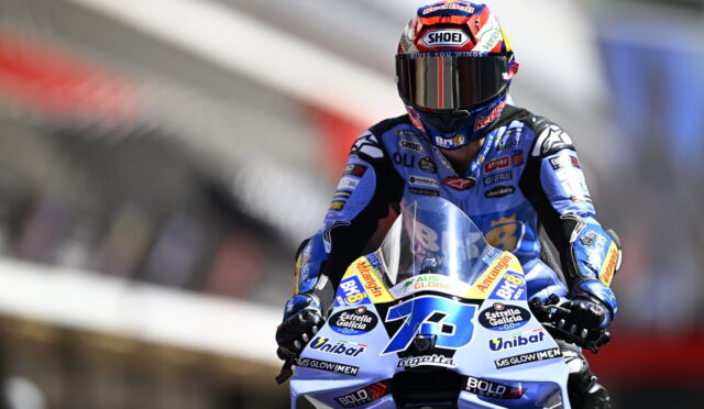 Motorcycle racer in a blue suit and red helmet speeds on a blue race bike at a track.
