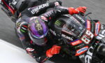 Racing motorcyclist in black and red leathers leans into a sharp turn on an Aprilia MotoGP bike with sponsor stickers visible, red gloves gripping the handlebars.