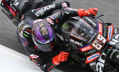 Racing motorcyclist in black and red leathers leans into a sharp turn on an Aprilia MotoGP bike with sponsor stickers visible, red gloves gripping the handlebars.