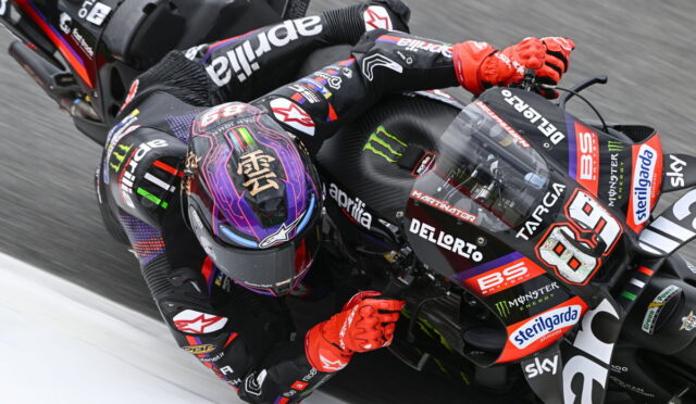 Racing motorcyclist in black and red leathers leans into a sharp turn on an Aprilia MotoGP bike with sponsor stickers visible, red gloves gripping the handlebars.