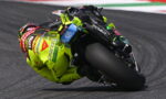 Moto racer in neon yellow suit leaning deep into a tight racetrack corner on a sportbike. The rider’s helmet and gear show sponsor logos typical of road racing.