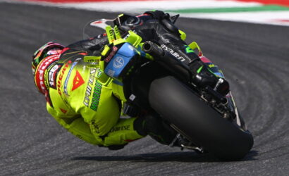 Moto racer in neon yellow suit leaning deep into a tight racetrack corner on a sportbike. The rider’s helmet and gear show sponsor logos typical of road racing.
