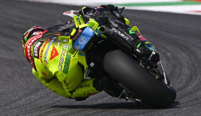 Moto racer in neon yellow suit leaning deep into a tight racetrack corner on a sportbike. The rider’s helmet and gear show sponsor logos typical of road racing.