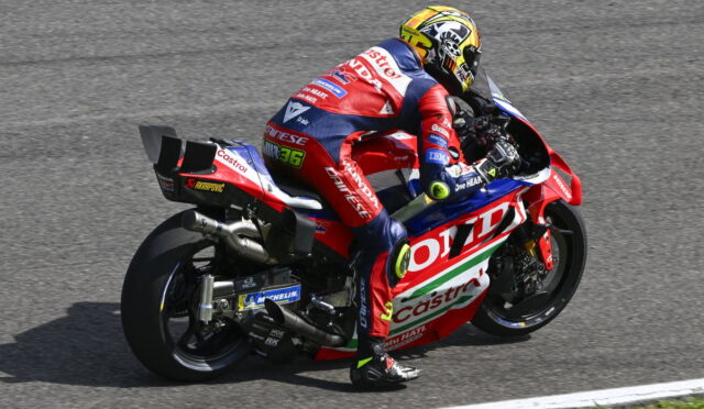 Professional motorcycle racer in a red, white, and blue suit leaning into a turn on a race track, helmeted and focused.