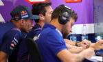 Three team members in blue uniforms wearing headsets lean over a desk, signing papers in a purple-branded room.