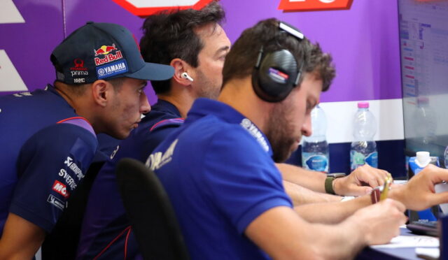 Three team members in blue uniforms wearing headsets lean over a desk, signing papers in a purple-branded room.